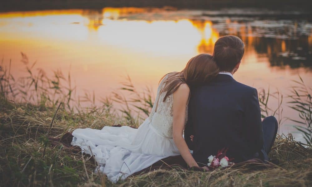 A couple of newly weds sit by the lake and watch the sunset.