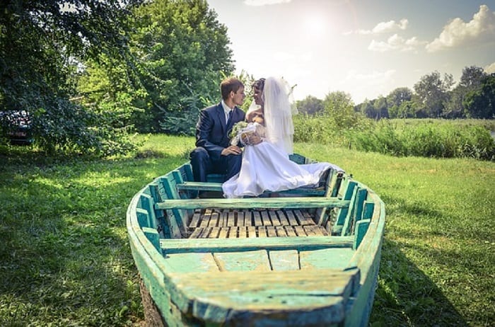 A recently married couple sit in a rustic looking rowing boat (turquoise) parked on dry land, surrounded by greenery. 