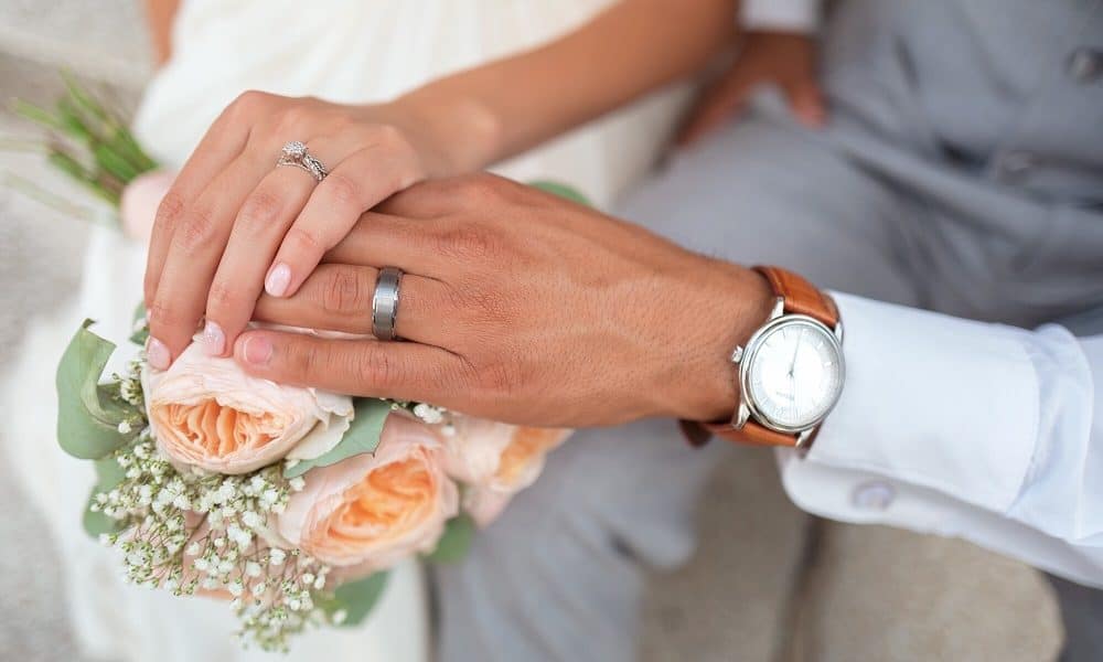 A recently married couple hold hands on top of a poesy of roses.