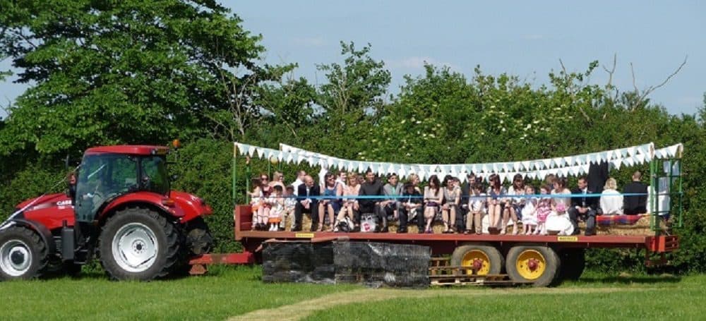 A tractor carrying a long trailer transporting a wedding party. Decorated with pastel colored bunting.